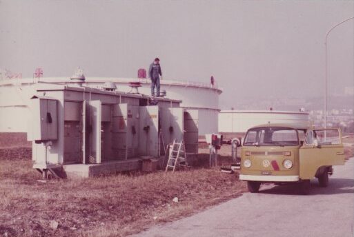 Ein Bild aus den frühen 80er Jahren mit einem VW-Bus vor Gasspeichern und Verteilerkästen, auf denen ein Techniker Rauchmelder installiert.