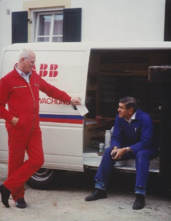 Vor einem Kleintransporter steht Franz Mittermeier in einem Roten Overall und spricht mit einem Mitarbeiter im Blaumann, der in der offenen Seitentür sitzt.