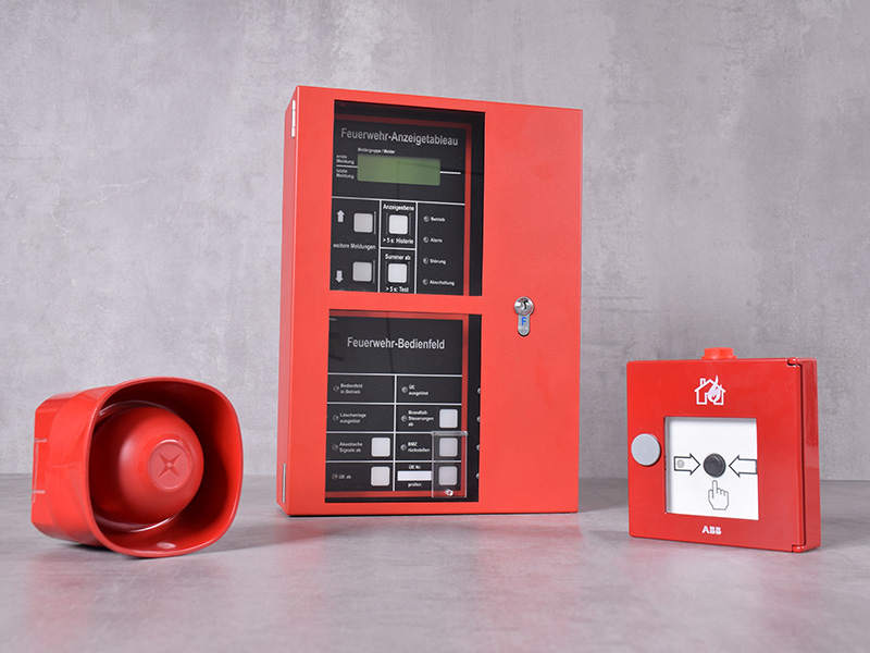 Ein Arrangement aus roten Brandmelde-Komponenten vor grauem Hintergrund: Sirene, Brandmeldezentrale und Notrufdrücker.