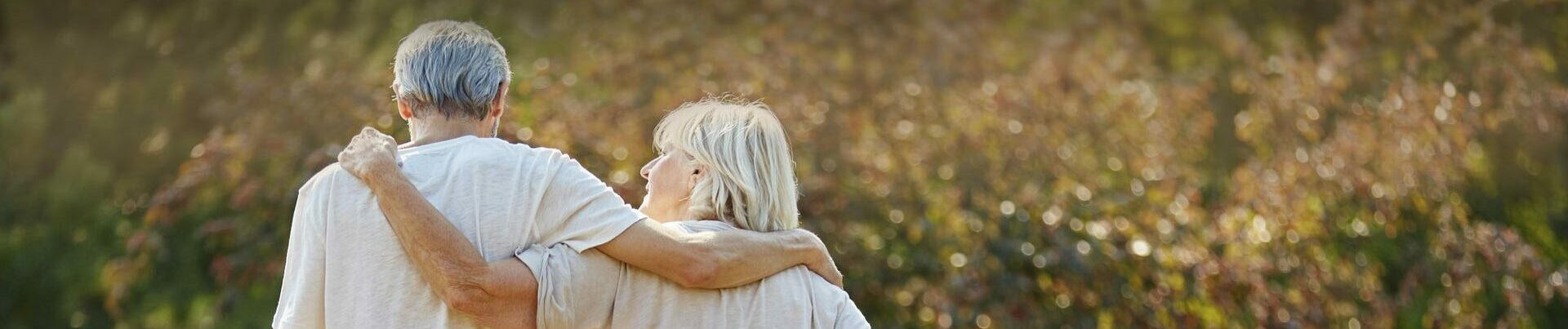 Ein älteres Paar in hellen T-Shirts spaziert Arm in Arm durch einen herbstlichen Garten